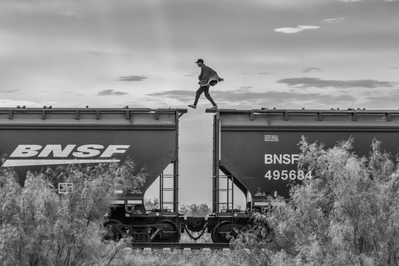 A person walks across the gap between two BNSF freight train cars, balancing carefully. The image is in black and white, with shrubs in the foreground and a cloudy sky above.