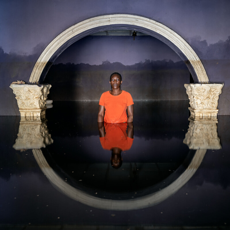 A person wearing an orange shirt stands waist-deep in reflective water, framed by a broken ornate arch with columns, against a purple wall. Their reflection is visible in the calm water below.