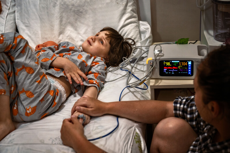A child in patterned pajamas lies in a hospital bed, holding an adult's hand. Medical monitoring equipment with visible vital signs is next to the bed. The child looks up, appearing thoughtful or anxious.