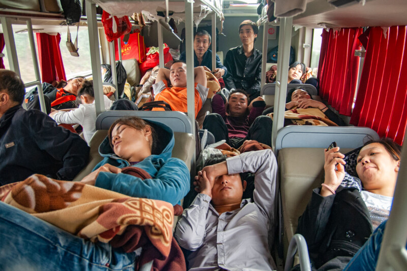 Several people lie on bunk beds and sit in an overcrowded sleeper bus, some sleeping, some using their phones. Red curtains and luggage are visible in the background. The overall mood appears cramped but relaxed.