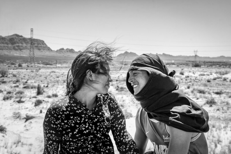 Two people stand close together in a desert landscape, facing each other and smiling. One wears a patterned shirt, the other has a shirt draped over their head. Mountains and power lines are visible in the background.