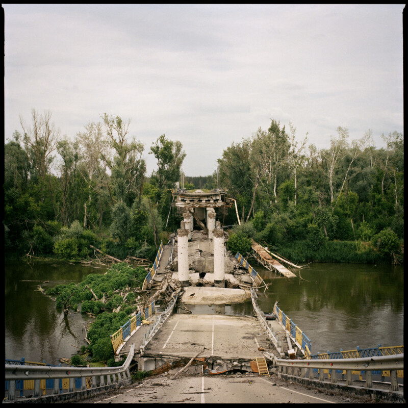 A damaged bridge with collapsed sections spans a river, surrounded by trees on both sides. Debris from the bridge lies in the water and on the roadway, making it impassable. The sky is overcast.