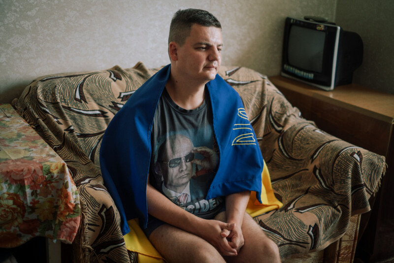 A man sits on a patterned couch with a Ukrainian flag draped around his shoulders, wearing a t-shirt with an image of a person and the word "PRESIDENT." An old television and a table with a floral tablecloth are nearby.