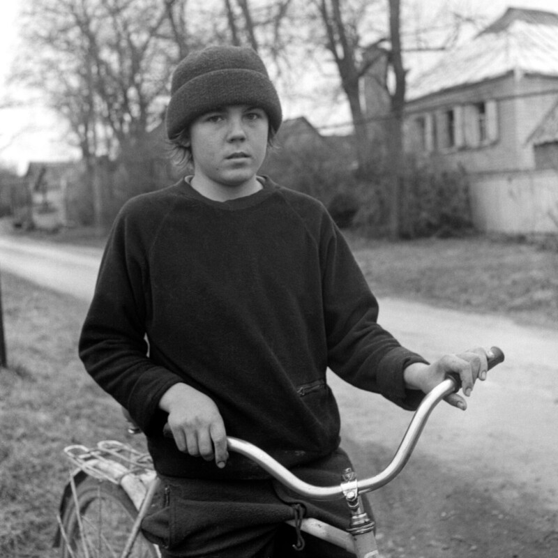 A young person in a knit hat and sweatshirt stands outdoors, holding the handlebars of a bicycle on a dirt road with trees and houses in the background.
