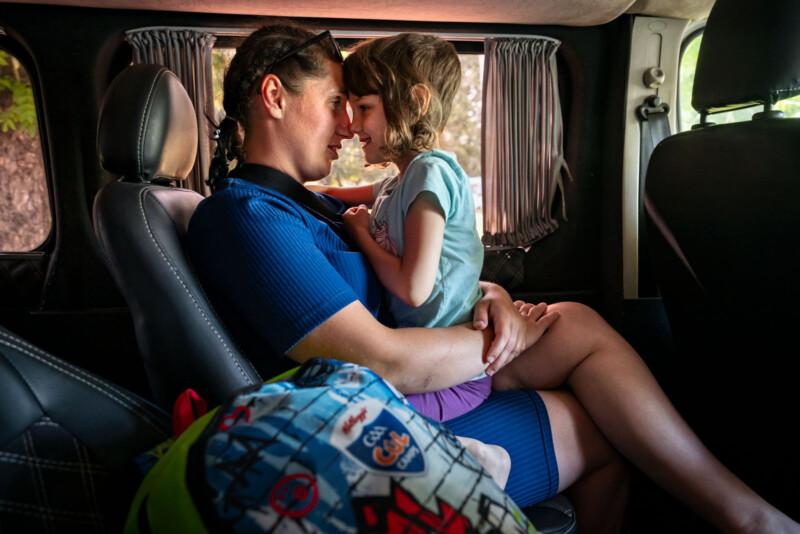 A woman and a young girl sit close together in the back seat of a car, touching foreheads and smiling warmly. A colorful backpack sits on the seat beside them. Light streams in through the window.