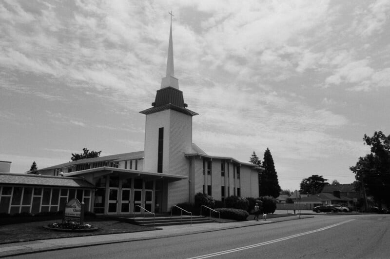 Black and white photo of a church with a tall steeple and cross, large windows, and an entrance facing a quiet street. A sign is visible near the entrance. Trees and clouds appear in the background.