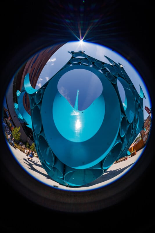 A fisheye lens photo shows a large, blue circular metal sculpture with multiple holes, set outdoors near buildings. The sun shines through the center, creating a bright spot and lens flare in a clear blue sky.