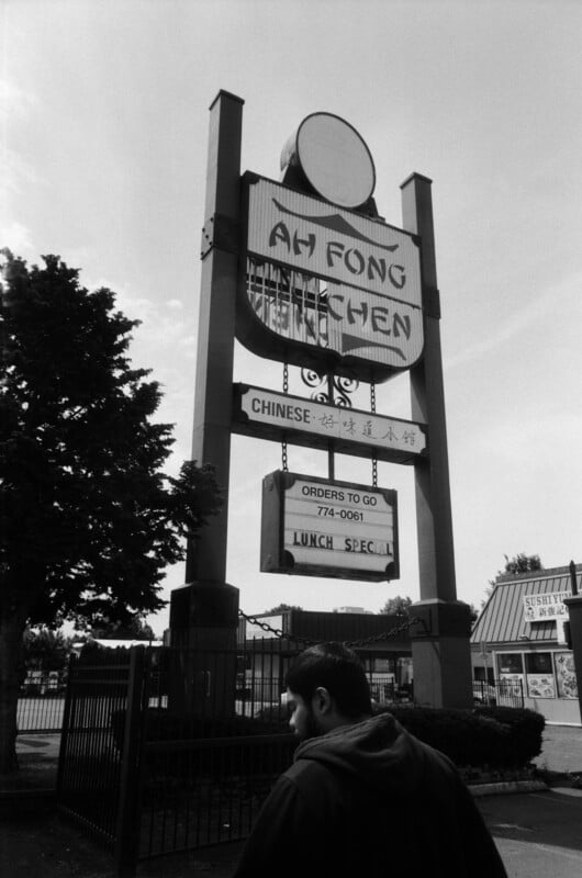 A man walks past a large roadside sign for "Ah Fong Kitchen," a Chinese restaurant, with trees and additional buildings visible in the background. The sign advertises orders to go and a lunch special.