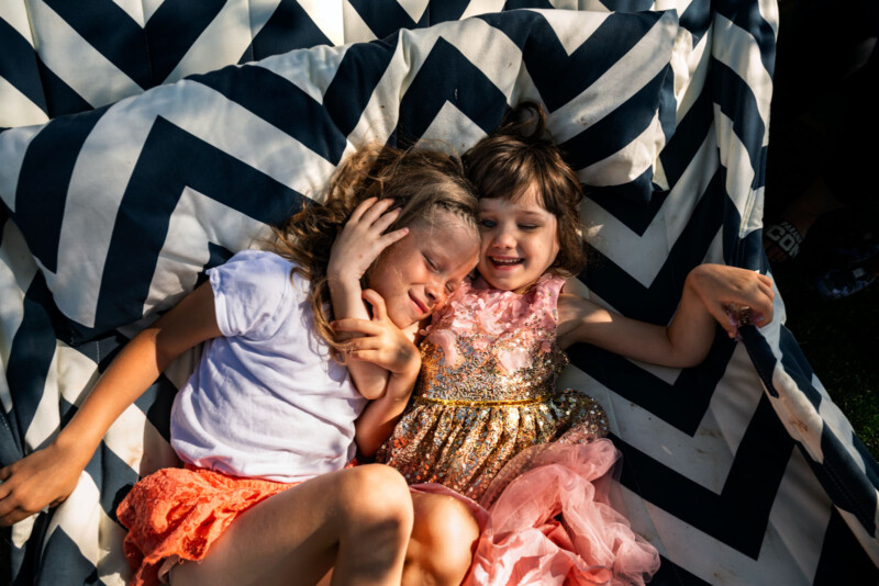 Two young girls laugh and hug while lying together on a black-and-white chevron patterned blanket outdoors, enjoying the sun. One wears a white shirt and orange shorts; the other wears a pink and gold dress.