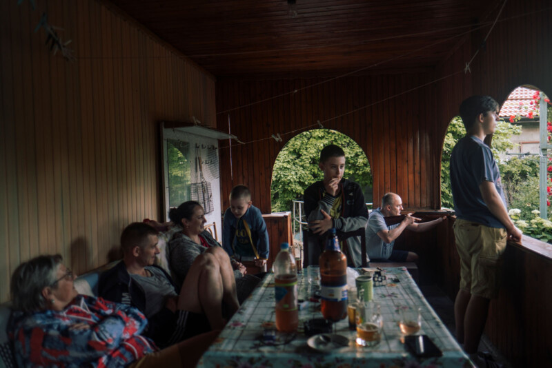 A group of people sit and stand on a covered porch around a table with bottles and glasses. Some are relaxing and talking, while one person looks outside over the railing. The porch is wooden and open to a green yard.