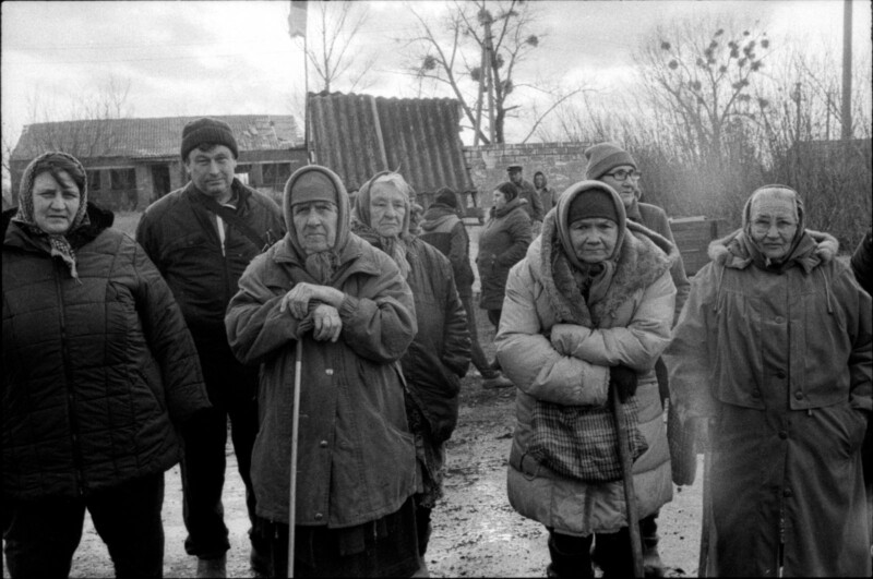 A group of elderly people, mostly women wearing winter coats and headscarves, stand outdoors on a muddy path. The background features leafless trees, old buildings, and cloudy skies.