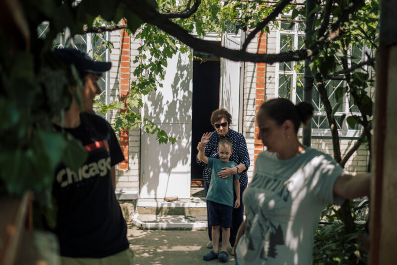 A woman waves from a doorway while standing with a young boy. Two other adults, a man and a woman, stand in the foreground near greenery, partially out of focus. The scene is set outside a house on a sunny day.