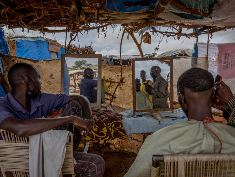A makeshift outdoor barbershop with two men seated in front of mirrors, one having his hair cut, as others watch. The area is covered by a roof of branches and tarps, with cloth and supplies scattered around.
