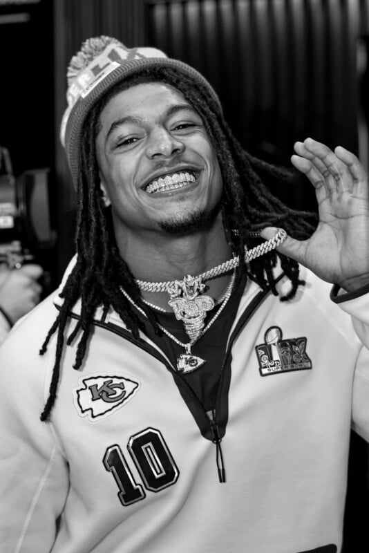 Man in a beanie and jacket smiles, showing off a large necklace with his hand. The jacket has a "10" patch and a football emblem. He has long braided hair and is standing against a striped backdrop. The image is in black and white.