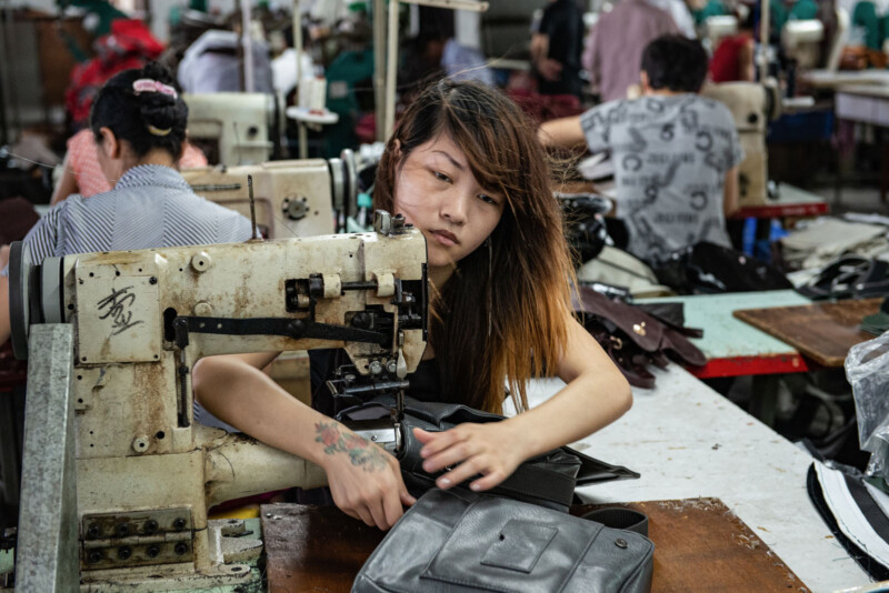 A woman with long hair operates an old sewing machine, stitching black fabric in a busy garment factory filled with other workers and sewing machines in the background.