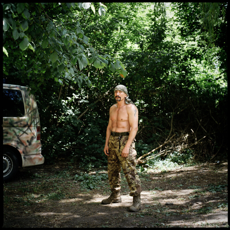 A muscular, shirtless man in camouflage pants and boots stands outdoors on dirt ground, surrounded by green trees and bushes, with a camouflage-painted van partially visible on the left.