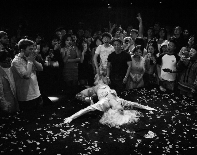A drag performer in a blonde wig and sparkly outfit lies dramatically on a confetti-strewn stage, surrounded by an enthusiastic crowd capturing the moment on their phones. The image is in black and white.