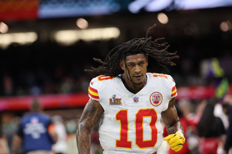 A football player in a white and red uniform and helmet with the number 10, runs on the field during a game. The player has long dreadlocks, and the background shows stadium seats and other players.