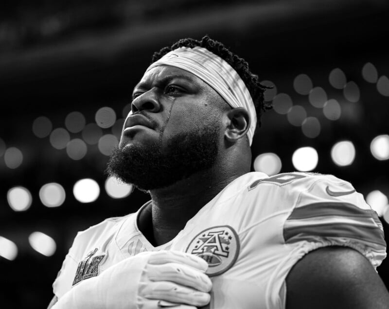 A football player in a uniform is standing with a hand on his chest, looking serious. He appears emotional, with a tear running down his cheek. The background is blurred with circular lights. The image is in black and white.
