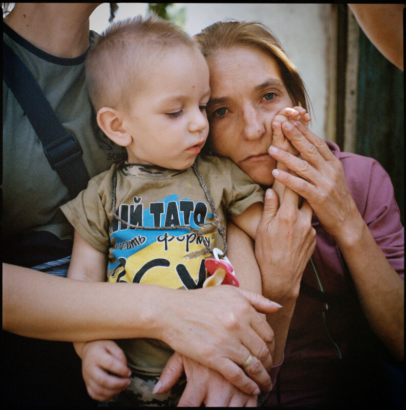 A woman holds a young boy on her lap, gently touching his hand to her face. Both appear thoughtful and close, with another adult partly visible, holding the boy for support. The mood is tender and intimate.