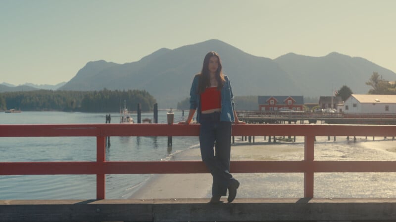 A woman in a red top and denim jacket leans on a red wooden railing by the water, with mountains and houses in the background under a clear sky.