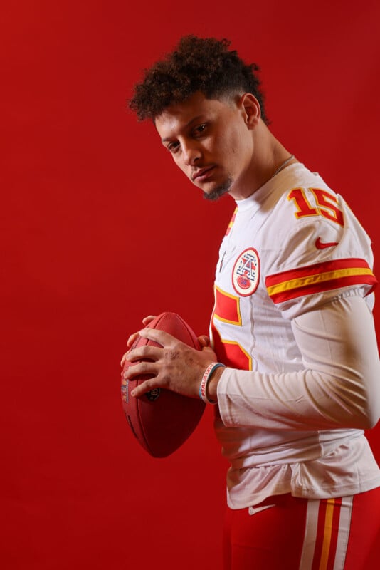 A football player in a white uniform with red and yellow accents stands against a red background, holding a football. He wears number 15 and has short curly hair. His expression is serious as he poses for the photo.