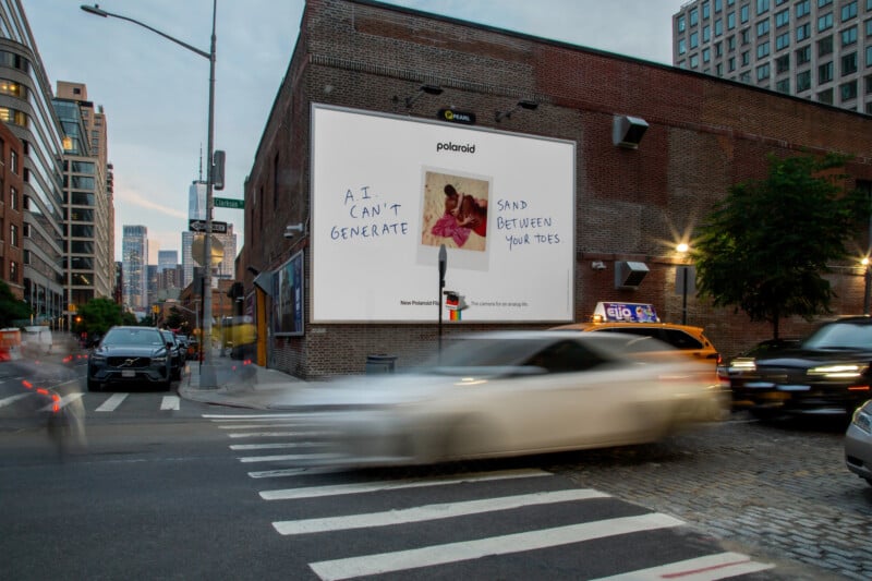 A large Polaroid billboard on a city street displays a photo of two people on a beach with handwritten text: "A.I. CAN'T GENERATE SAND BETWEEN YOUR TOES." Cars drive by, with buildings in the background.