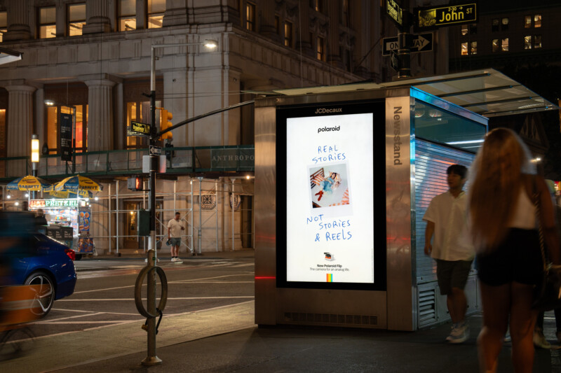 A digital billboard at a city bus stop displays a Polaroid advertisement reading, "Real stories, not stories & reels," with a hand holding a Polaroid photo. People and cars move around in the evening city scene.