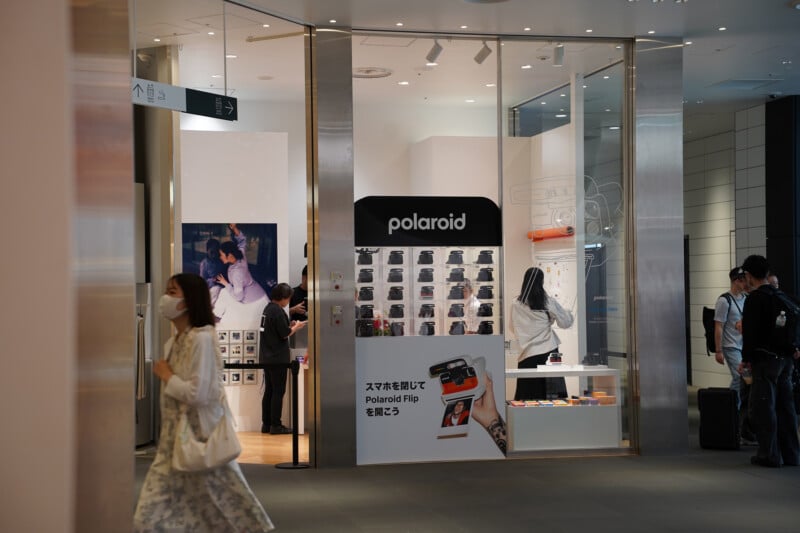 A Polaroid store in a mall with large glass windows, Polaroid cameras on display, and people walking by. A woman in a mask and dress walks past, while others browse inside the brightly lit store.