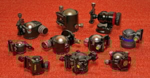 Various black tripod ball heads with adjustment knobs and level indicators are arranged on a textured red surface. They vary in size and design, showcasing different features and mechanisms.