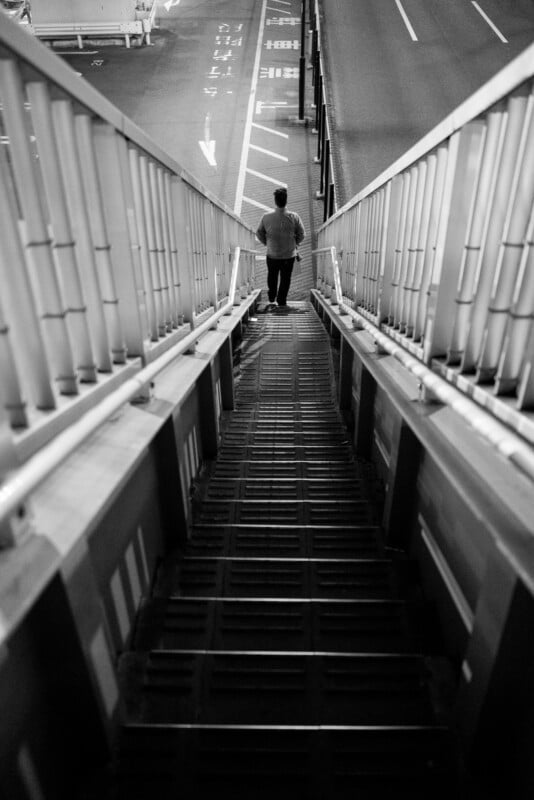 A person in a light sweater is walking down a long, steep staircase in an urban setting. The stairs are bordered by railings and lead to a street with visible lane markings. The image is in black and white.