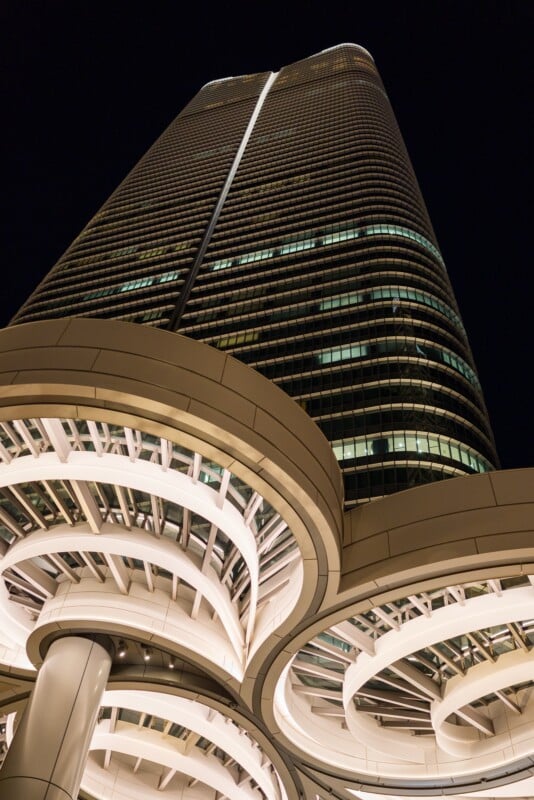 Upward view of a modern skyscraper at night, with circular architectural elements at the base. The building is illuminated, highlighting its tall, sleek design against the dark sky.