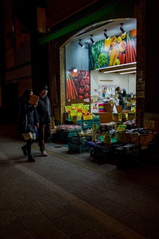 A night street view of two people walking past a brightly lit produce shop. The store has vegetables and fruits displayed outside in crates, with a worker visible inside.
