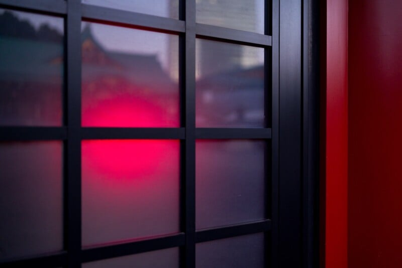 Frosted glass window with a black grid frame, showing a soft, diffused red light shining through. The red light creates a warm glow, enhancing the surrounding dark and neutral tones. A hint of a structure is visible beyond the glass.