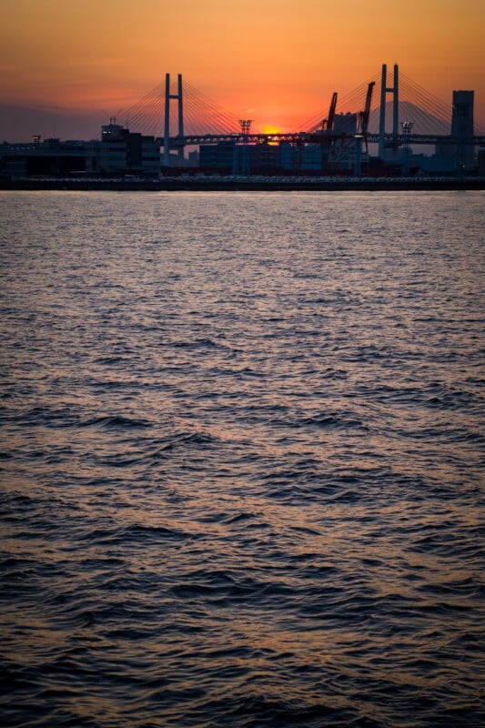 Sunset over a calm, rippling body of water with a silhouetted cable-stayed bridge and industrial structures in the distance. The sky is painted in warm orange and purple hues, with the sun partially hidden behind the bridge.