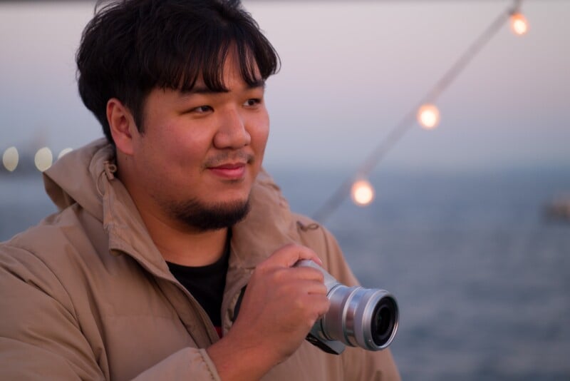 A person with short dark hair and a beard holds a camera, standing outdoors near a body of water. The individual is wearing a beige jacket. The background is softly lit with string lights and an evening sky.