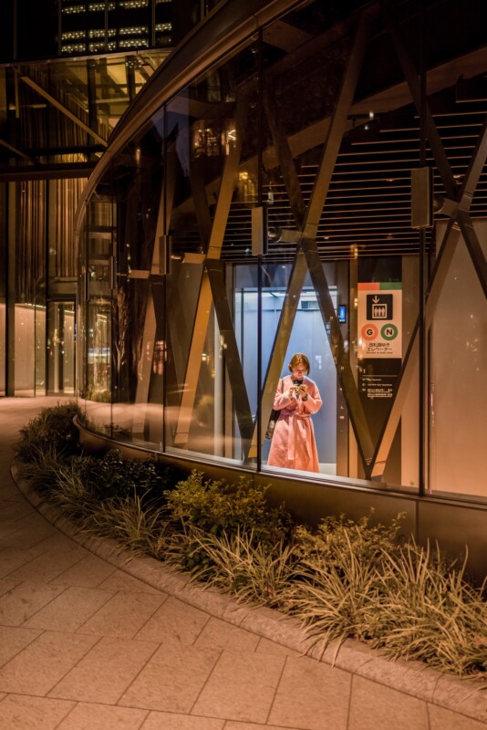 A person in a pink coat stands in a well-lit glass elevator at night, holding a phone. The surrounding area has modern architecture with large glass panels and geometric patterns, and some plants are visible by the walkway.