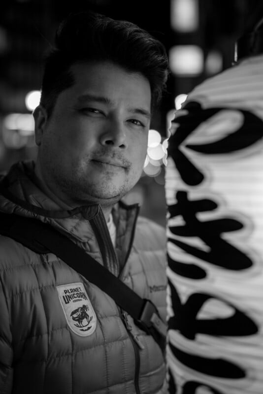 A person with short hair stands beside a large lantern with bold, vertical writing. They wear a puffy jacket with a patch on it. The background is softly blurred with circular lights, suggesting a nighttime setting in a city. The image is in black and white.