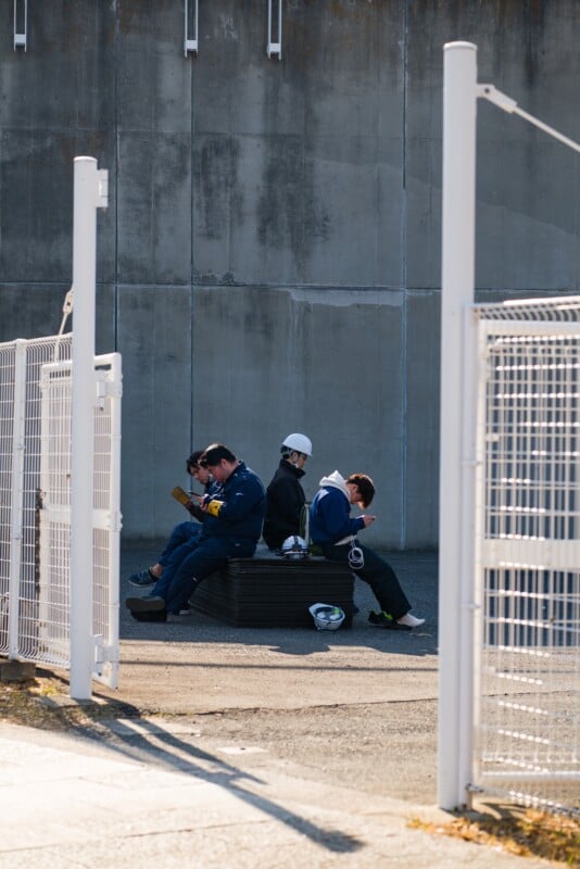 Four construction workers sit on a black bench, taking a break. One wears a white hard hat, while others are in uniform, using phones or reading. They are beside a tall concrete wall, framed by white fence posts.