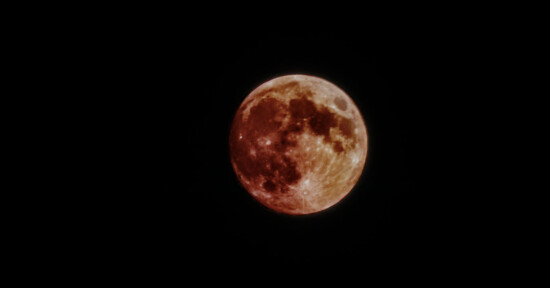A full moon with a reddish hue is set against a dark sky. The surface of the moon is detailed, showing lunar craters and shadow contrasts. The reddish tint suggests a possible lunar eclipse or atmospheric effect.