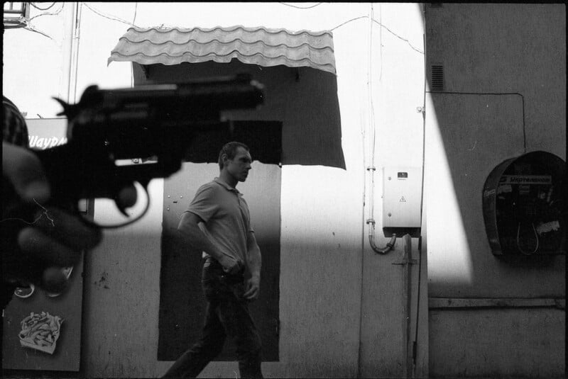 A man walks past a building wall while a hand points a gun in the foreground. The scene is in black and white, with strong shadows and an old-fashioned phone booth visible on the right.