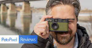 A person holding a green film camera up to their eye, smiling, with a bridge and water in the background. The image includes text reading "PetaPixel Reviews" at the bottom left corner.