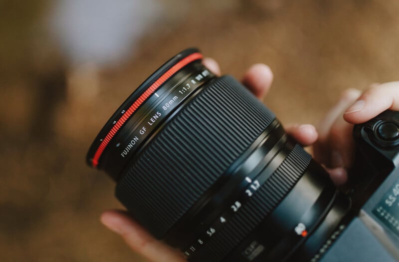 A close-up of a person holding a camera with a FUJINON GF Lens 80mm f/1.7, featuring a prominent red ring and textured focus ring, with a blurred brown background.