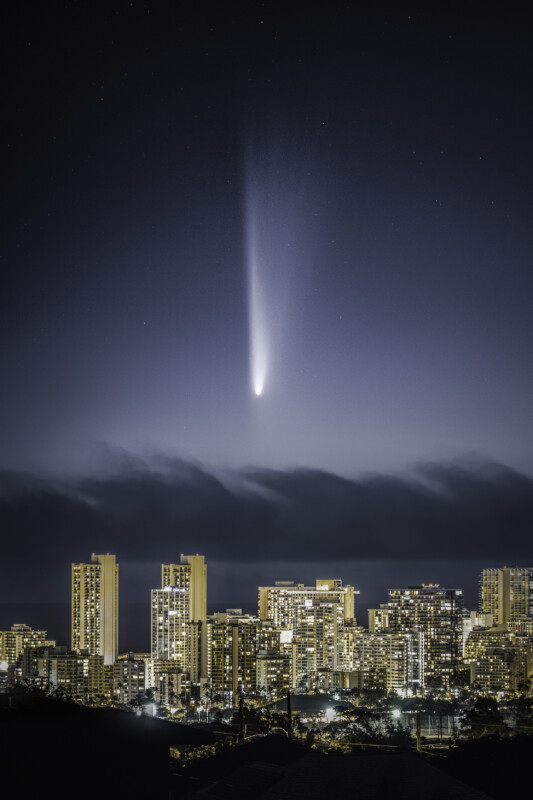 A bright comet with a long glowing tail streaks across the night sky above a city skyline with illuminated high-rise buildings. Thin clouds hover above the horizon, and stars are visible in the sky.