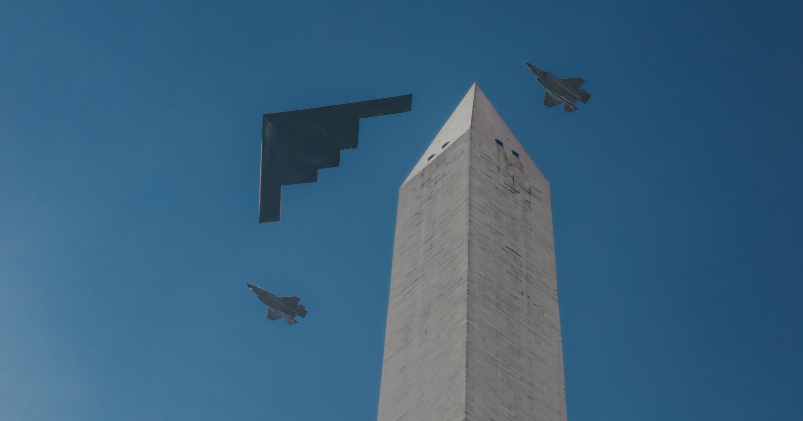 Three military aircraft fly past the Washington Monument against a clear blue sky—two fighter jets flank a stealth bomber in a formation above the monument's pointed tip.