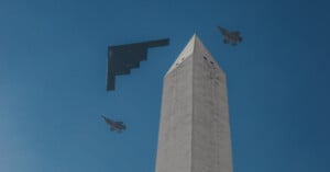 Three military aircraft fly past the Washington Monument against a clear blue sky—two fighter jets flank a stealth bomber in a formation above the monument's pointed tip.