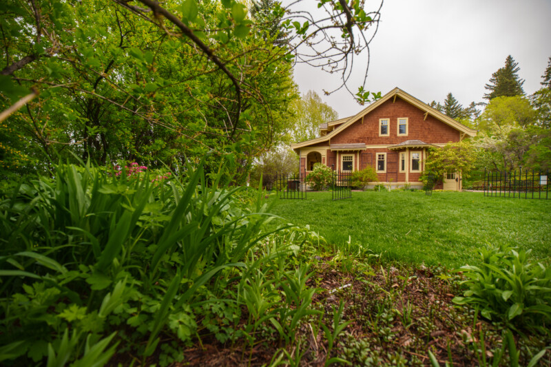 A charming red house with a front porch sits amid lush green grass, trees, and garden plants on a cloudy day, creating a peaceful, welcoming atmosphere.
