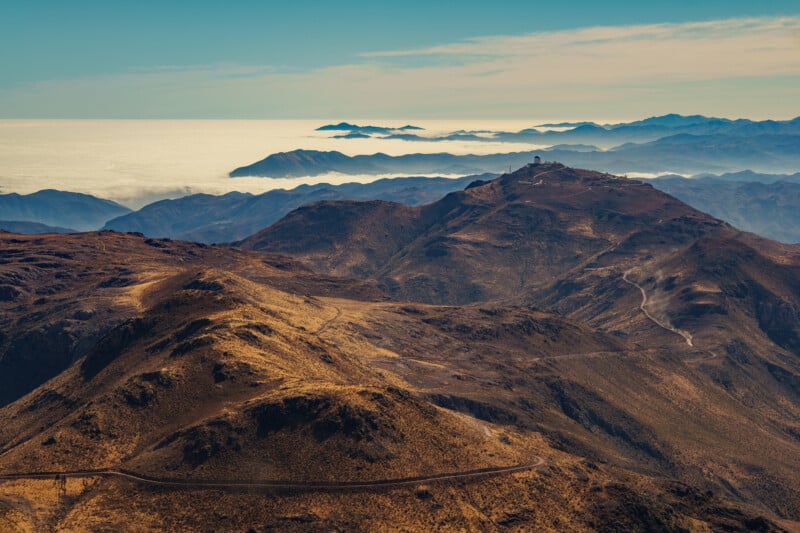 Mountain ranges under a clear sky, with observatories visible on a distant ridge and layers of hills fading into mist; a winding road crosses the dry, golden landscape.