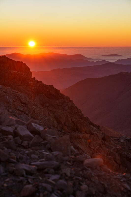 The sun sets over a rugged mountain landscape, casting an orange and purple glow across rocky slopes and distant ridges under a clear sky.