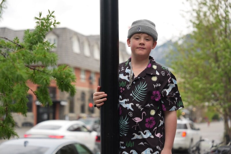 A boy wearing a gray beanie and a black shirt with colorful dinosaur prints stands outdoors, holding onto a black pole. Trees, buildings, and parked cars are visible in the background.